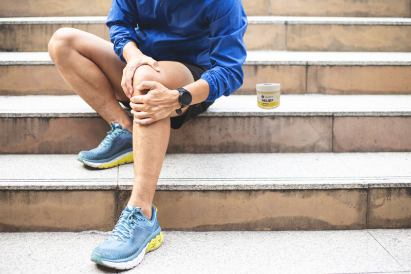 Athlete sitting on steps, holding their knee in recovery, with Cannafyl Relief CBD Salve nearby for muscle soreness relief.